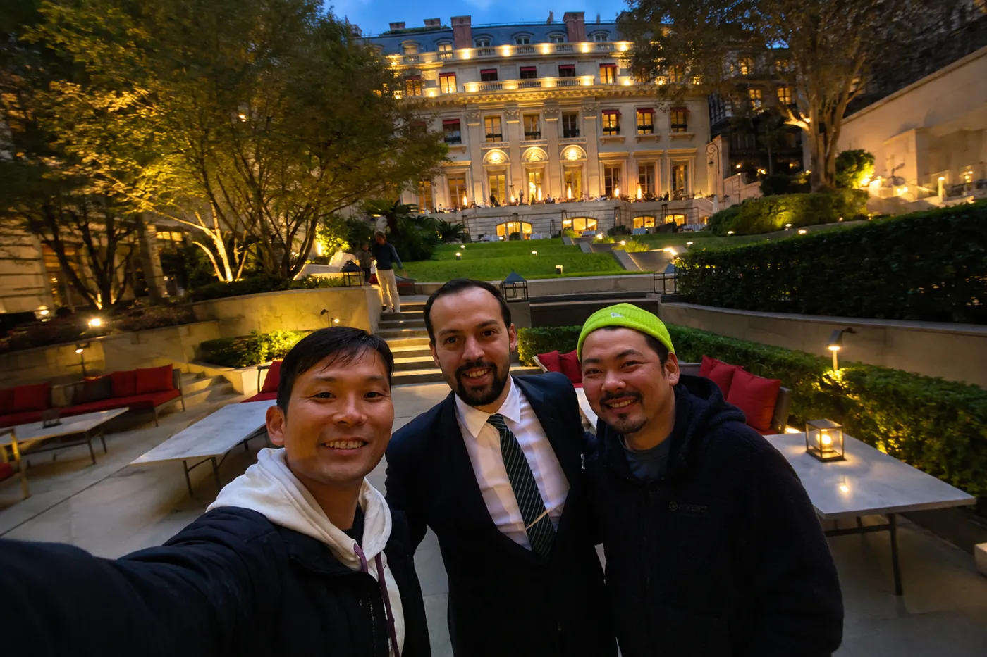A garden at night. Three people standing together in front of a lit building, a man in a suit between them