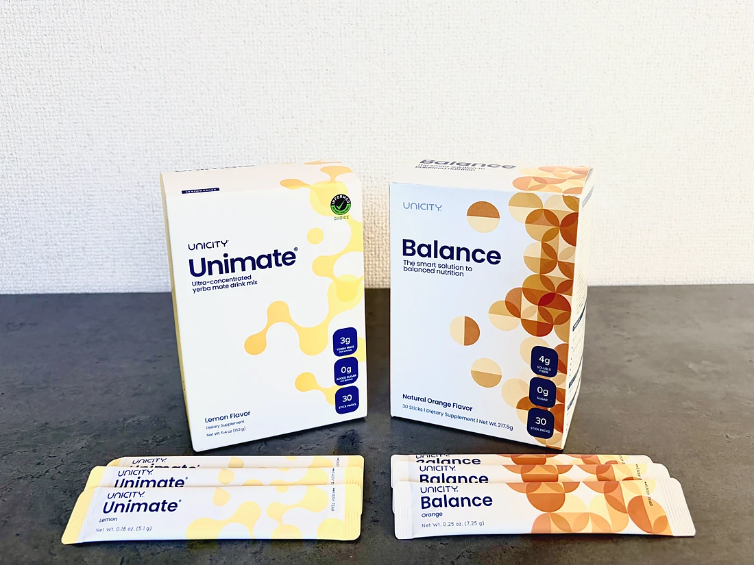 Cajas de Unimate y Balance apoyadas en una pared blanca, con sobres de cada uno al frente