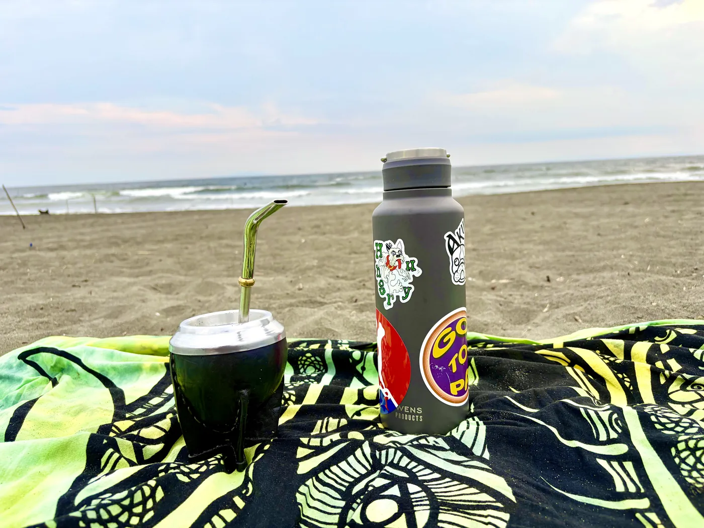 夕暮れの砂浜。緑と黄色の模様の布の上に、革を巻いた黒いマテ壺とボンビーシャ、ステッカーの貼られた水筒。奥に波打ち際と空