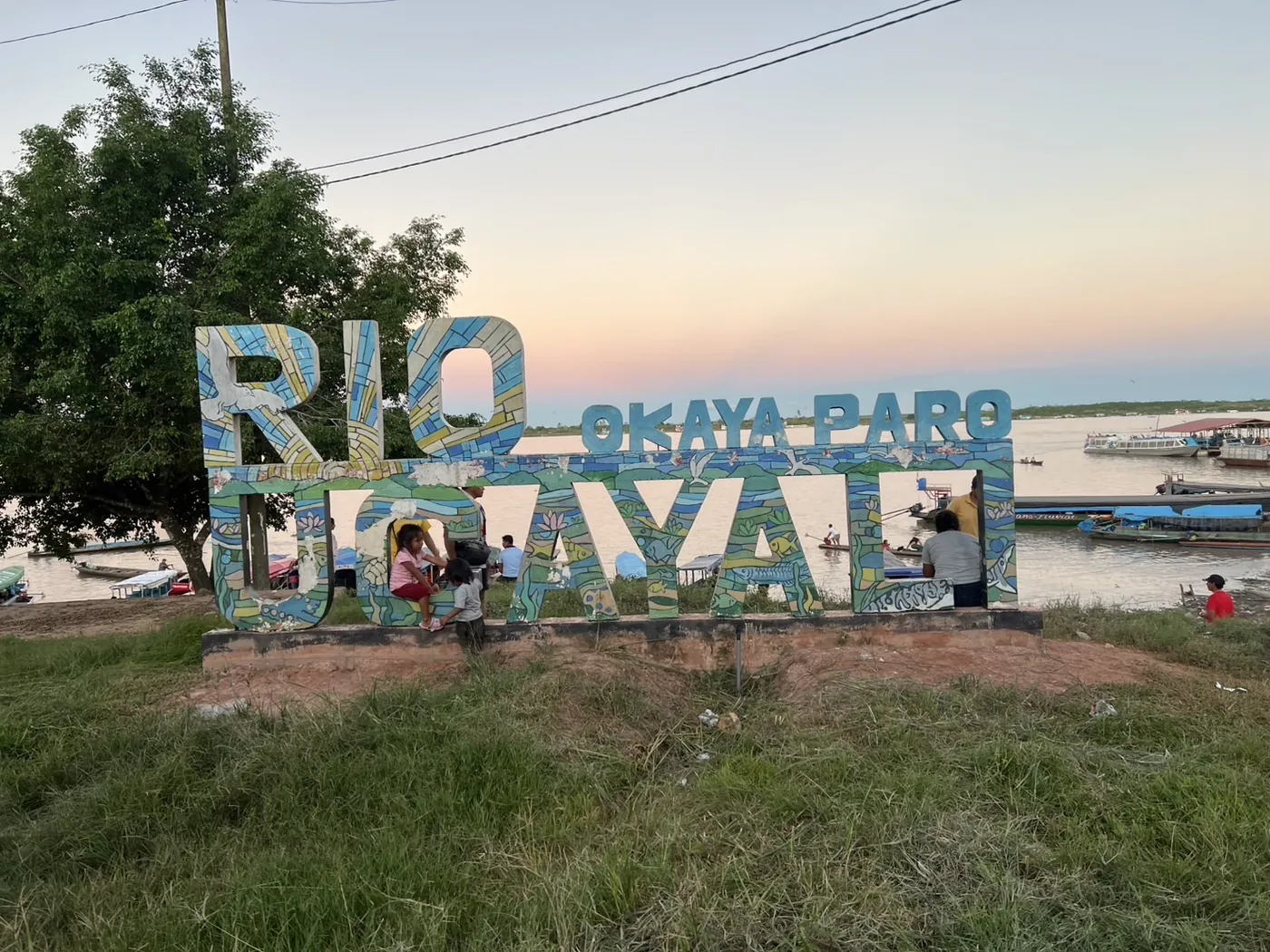 夕暮れの川辺。「RIO UCAYALI」と大きく描かれた立体文字のモニュメント。奥に川と停泊する舟が見えている