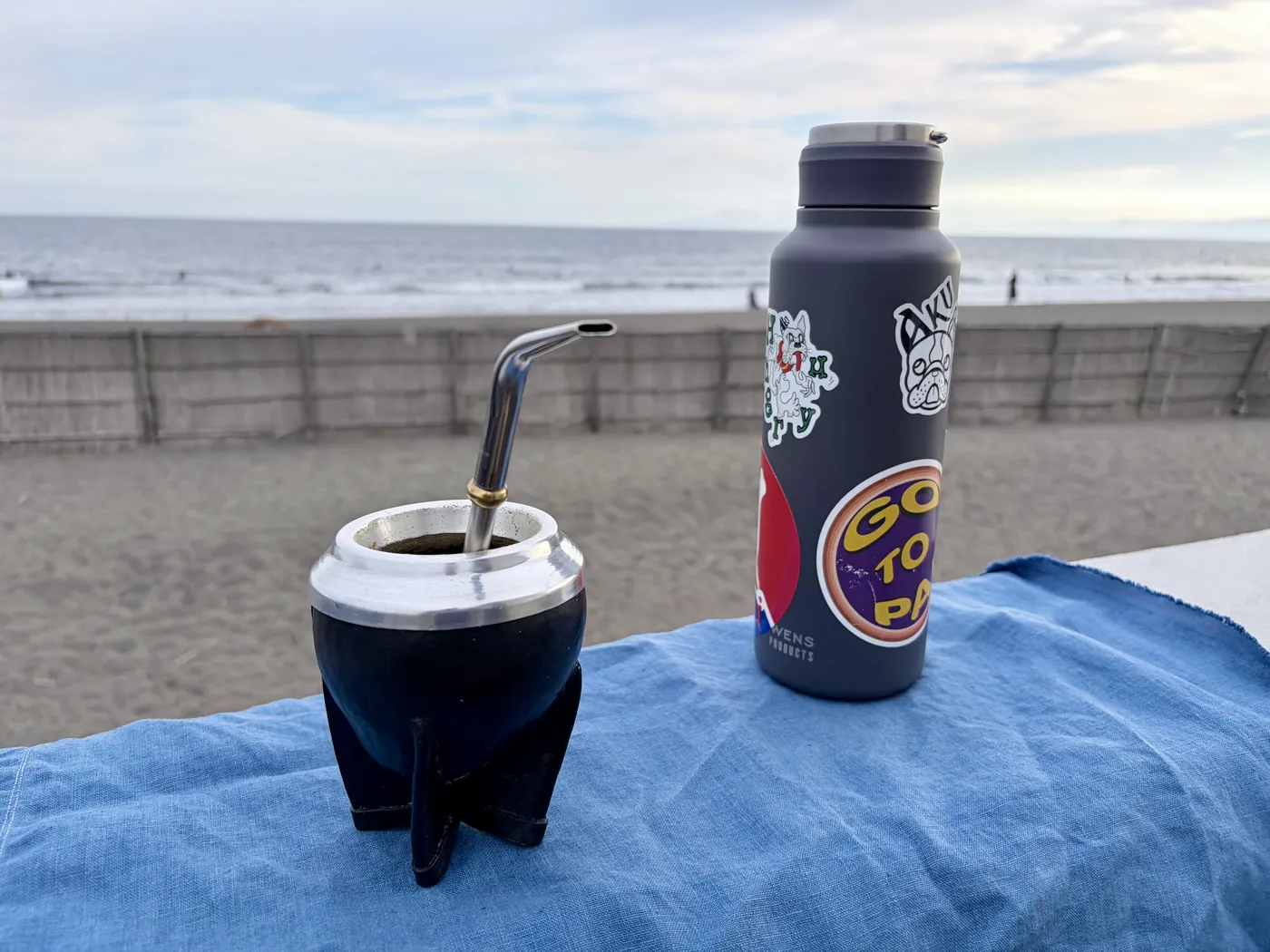 砂浜に敷いた青い布の上に置かれた、ひょうたん製のマテ壺とボンビーシャ。隣にステッカーの貼られた水筒。奥に波打ち際と海
