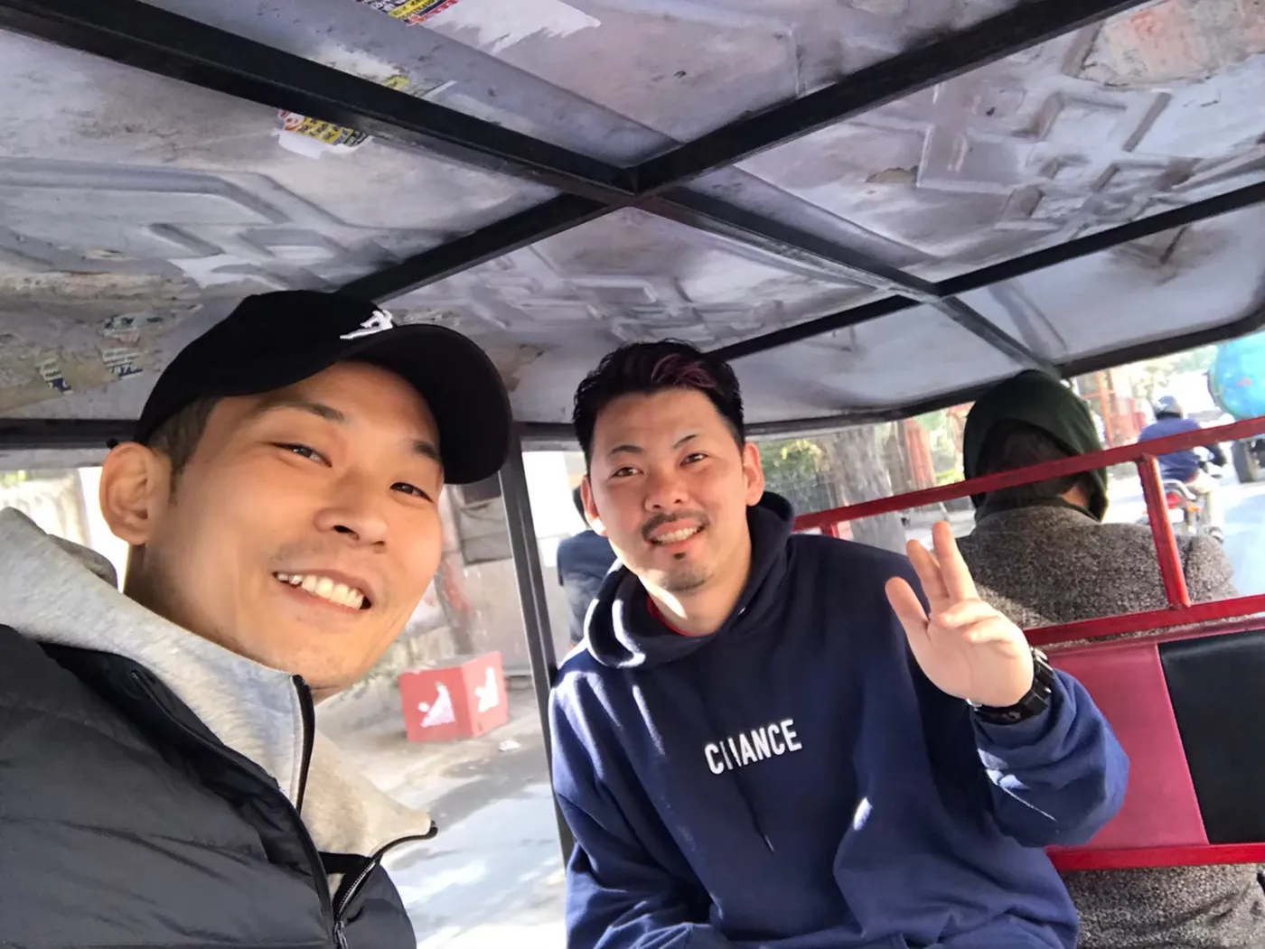 兩個男人並排坐在人力三輪車的後座，一個戴黑色帽子，另一個比出勝利手勢